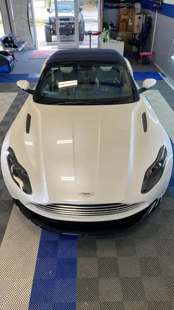 A white Aston Martin convertible with a black roof parked inside an automotive shop, viewed from above the front. Another vehicle is visible through the glass entrance.