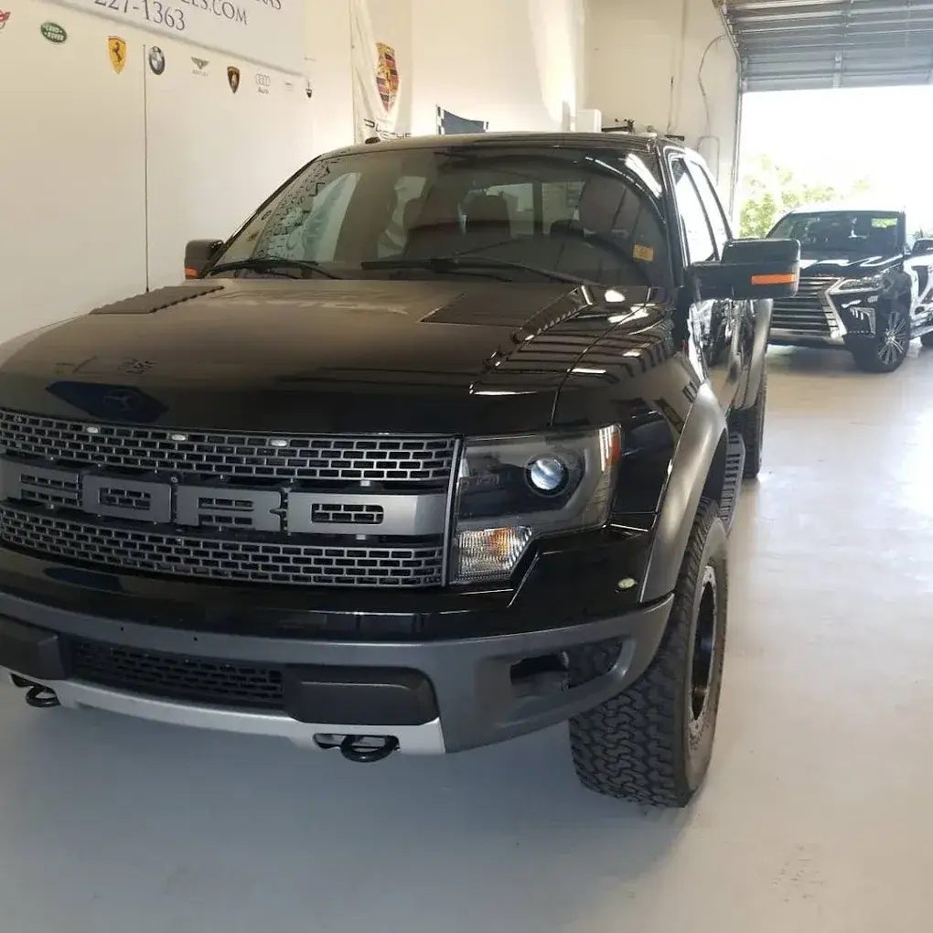 A black Ford pickup parked inside an automotive garage, with a black Lexus SUV behind it near an open bay door. Automotive logos decorate the wall.