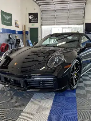A glossy black Porsche convertible parked inside an automotive workshop on a gray, black, and blue checkered floor beneath a raised garage door.