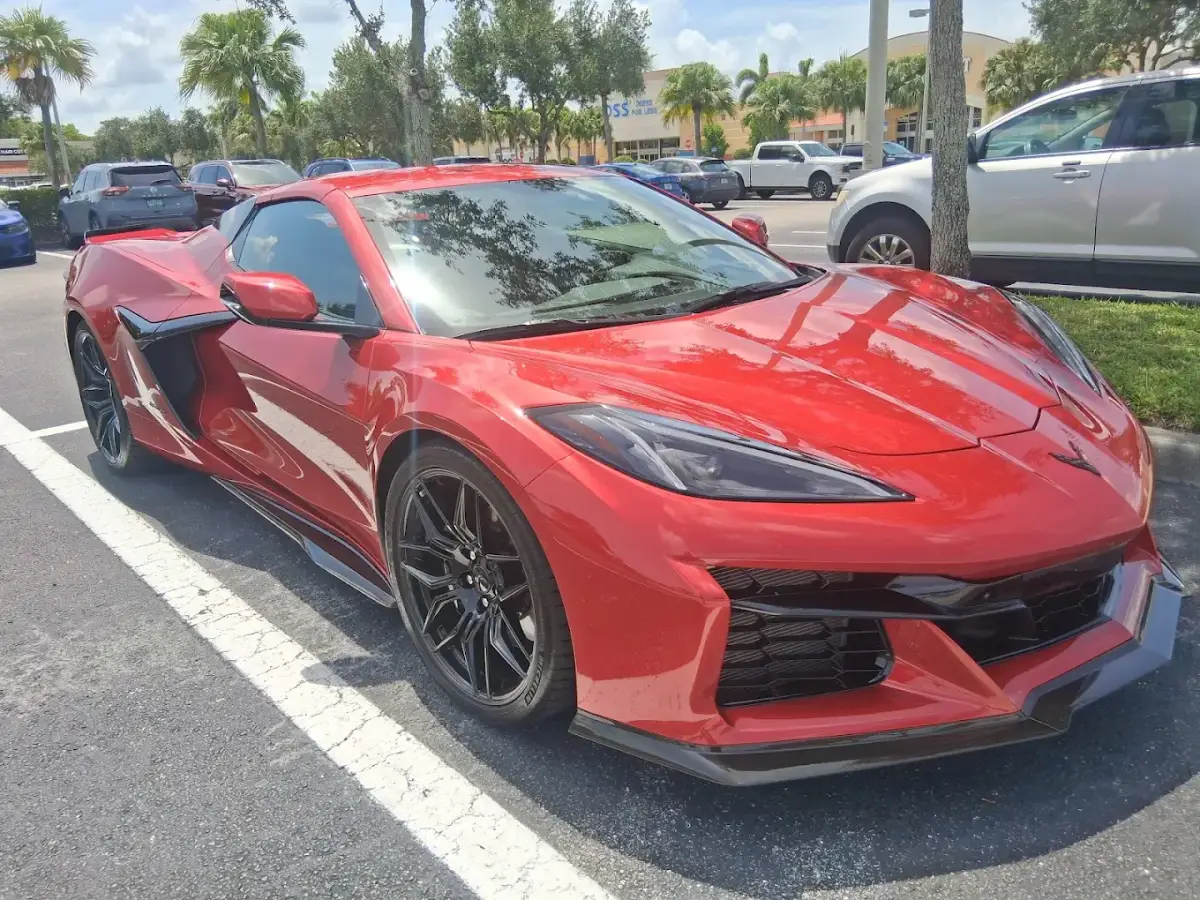A glossy red Chevrolet Corvette with black wheels parked outside a shopping center, viewed from the front corner.