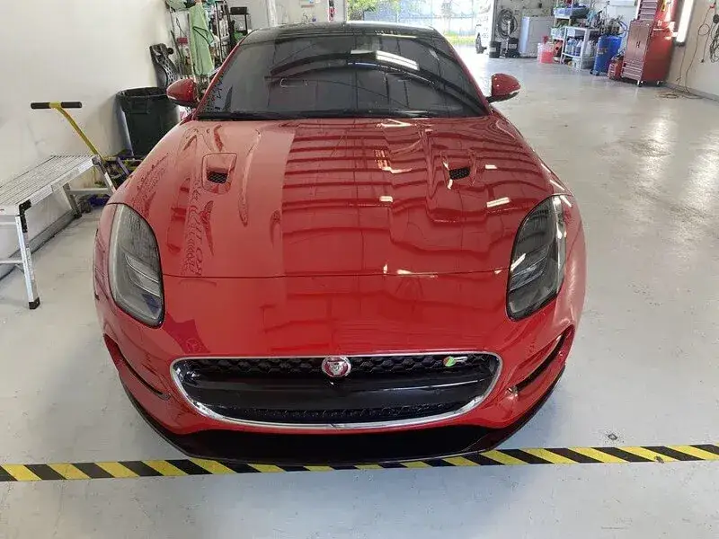 Front view of a glossy red Jaguar sports car inside a workshop, with tools, shelving, and cabinets around it.
