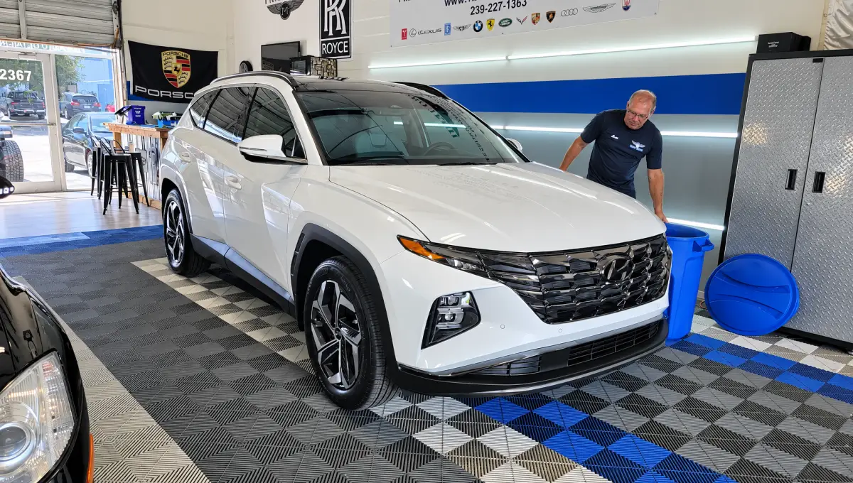 A man stands beside a white Hyundai SUV inside an automotive shop with bright wall lights, patterned flooring, automotive banners, and metal storage cabinets.