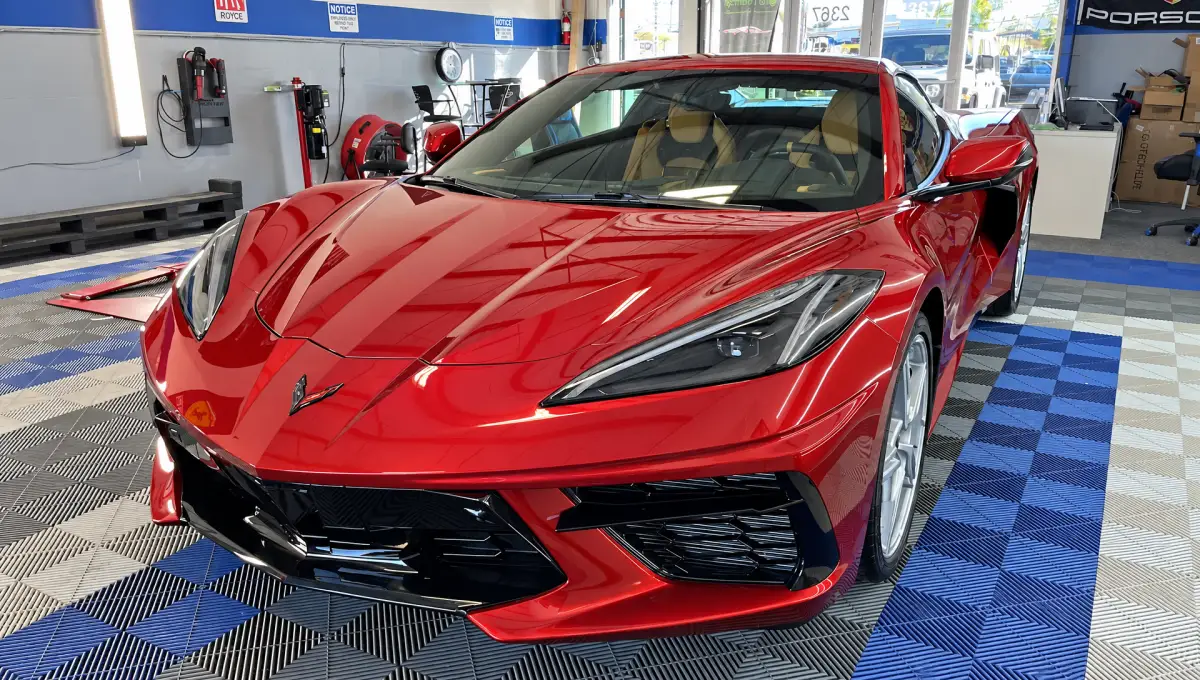 A glossy red Chevrolet Corvette parked inside an automotive workshop, with a blue, gray and black tiled floor and equipment along the walls.