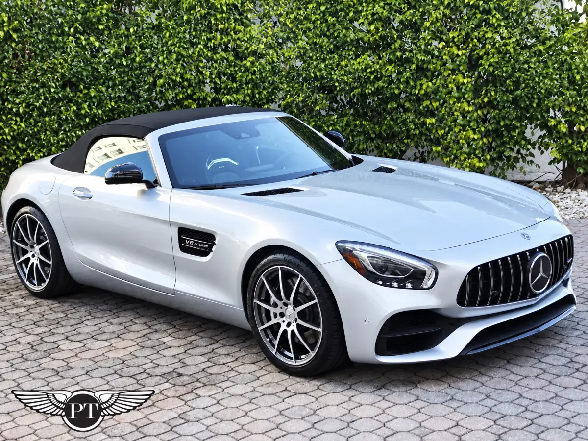 A silver Mercedes-Benz convertible with a raised black soft top sits on a paved driveway in front of a hedge. A winged PT logo appears at the bottom left.