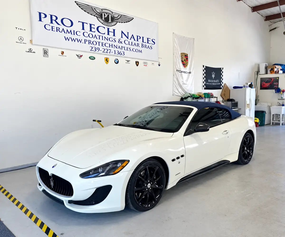 A white Maserati convertible with a dark soft top and black wheels sits inside a workshop beneath a Pro Tech Naples sign advertising ceramic coatings and clear bras.