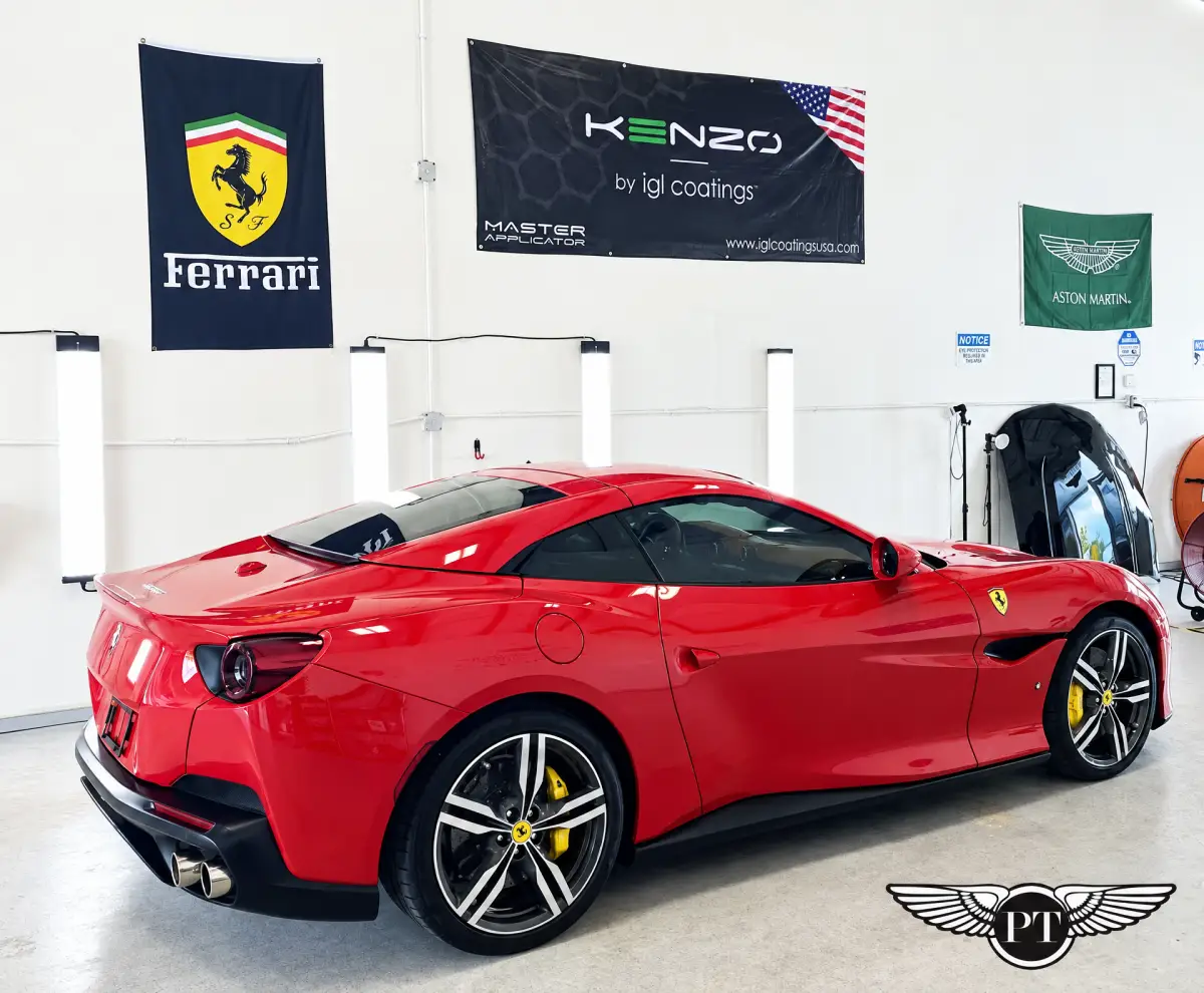 A glossy red Ferrari parked inside a brightly lit automotive workshop, with Ferrari, Aston Martin and vehicle-coating banners on the wall.