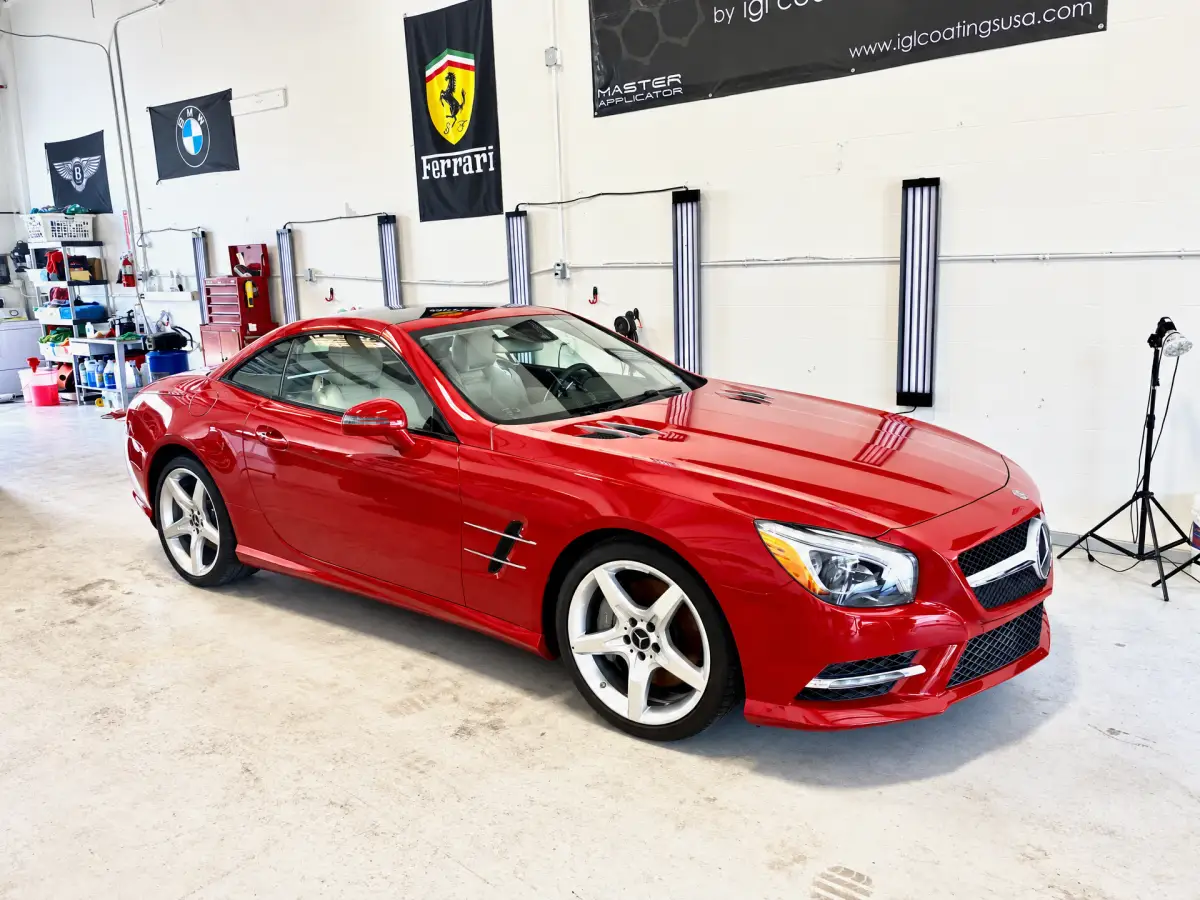 A glossy red Mercedes-Benz parked inside an automotive workshop with wall-mounted lights, detailing supplies, and automotive banners. No people are visible.