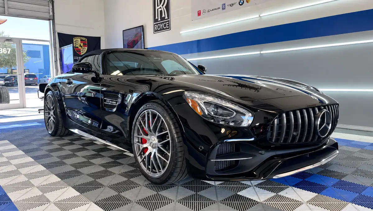 A glossy black Mercedes-Benz sports car parked inside a garage with bright wall lights, automotive banners, and a checkered floor.
